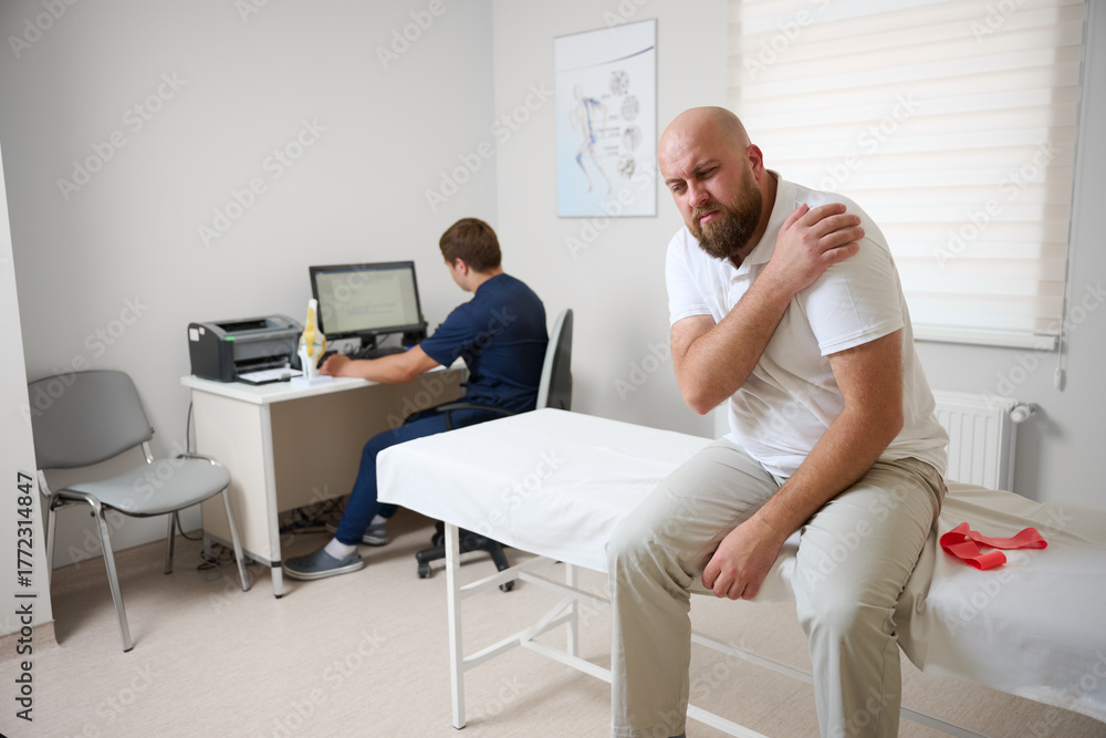 Obraz premium Male patient being examined in clinic for shoulder pain. Male suffering from shoulder pain. Patient sitting on examination couch holding his arm while doctor works at computer in background.