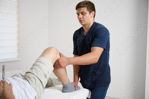 Physiotherapist examining patient knee joint during rehabilitation therapy session in modern medical clinic. Man receiving orthopedic treatment and physical therapy for injury recovery.