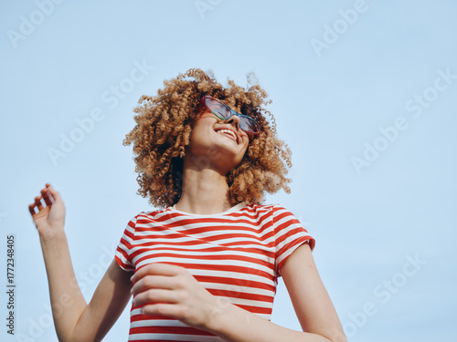 Young person with curly hair smiles under a clear blue sky, wearing a red and white striped shirt and sunglasses, radiating carefree summer energy and playful confidence.