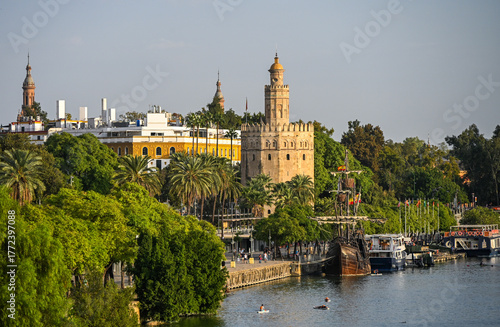 View of the Torre del Oro beside the Guadalquivir River in Seville, surrounded by palm trees, boats, and historic architecture. The golden tower glows in the afternoon light