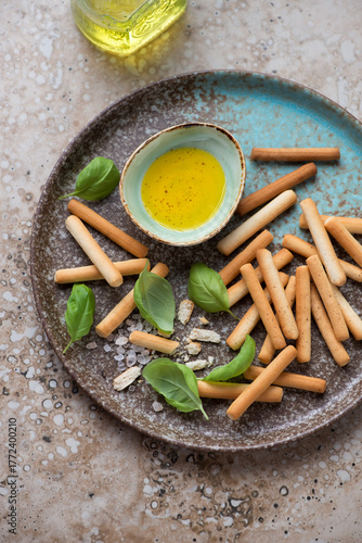 Brown plate with salted breadsticks, olive oil and green basil, vertical shot on a beige granite surface, high angle view