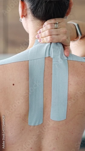 Young woman with kinesiology tape on her neck and shoulder gently massaging the area with her hand. Concept of muscle recovery, pain relief and self care using therapeutic taping techniques