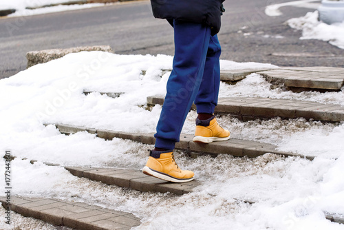 Man in winter boots walking carefully on icy snow covered steps outdoors, slippery conditions, risk of falling during winter season. Close-up of male legs in winter shoes stepping on frozen staircase