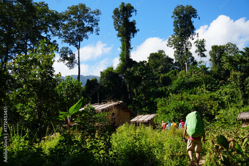 Fototapeta premium Adventurous group exploring lush jungle trail with traditional village huts, vibrant colors, and sunny skies, perfect for travel blogs and eco-tourism ads