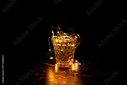 Guirlande lumineuse scintillante dans un verre transparent avec reflets dorés, verre vue de face centré dans l'image et posé sur un fond brun foncé avec étoiles dorées brillantes parsemées autour