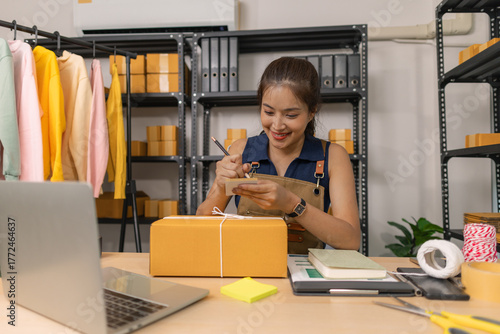 Young female entrepreneur writing a shipping address on a label for a parcel, preparing a package for delivery at her home office. She manages an online business and organizes shipments to customers.