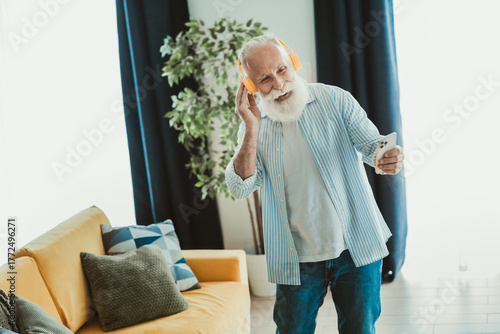 Happy senior man with white beard listens to music on bright orange headphones while holding a smartphone at home smiling