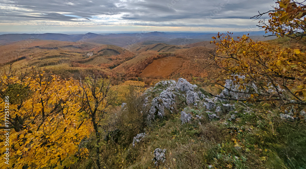 Obraz premium Vivid autumn panorama from Három-kő peak in the Bükk Mountains, Hungary. Rolling forested hills painted in gold, orange and green beneath a dramatic cloudy sky.
