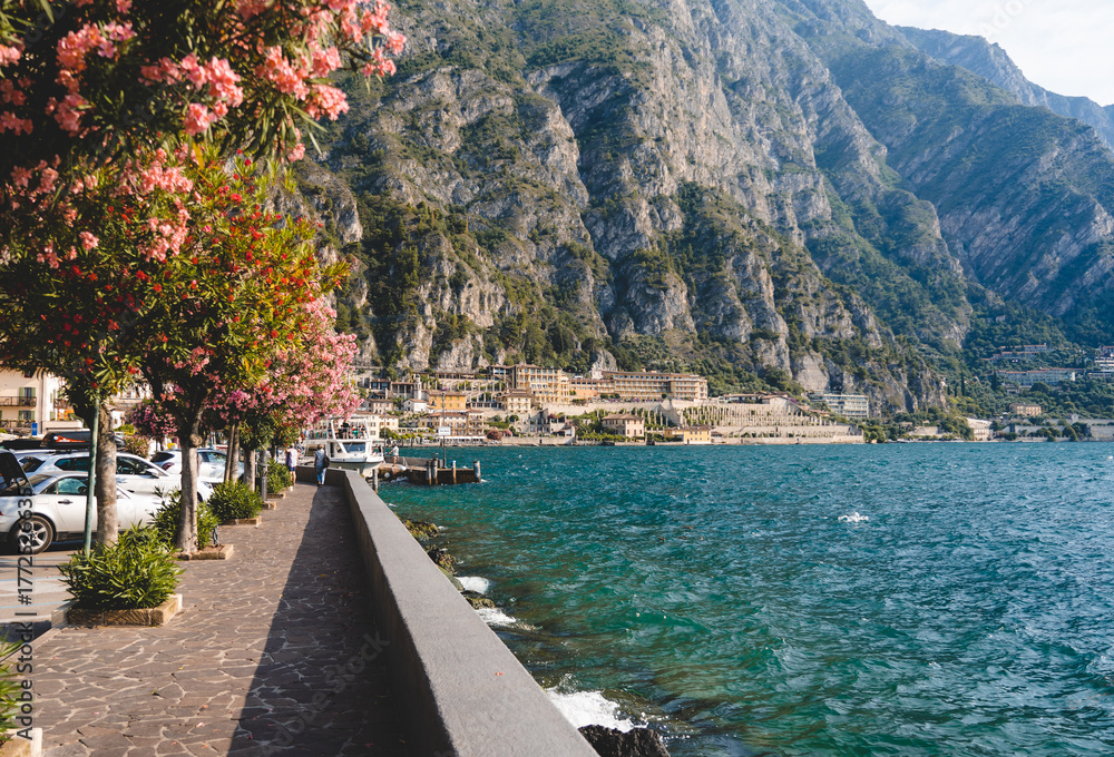 Obraz premium Scenic promenade with pink blooming trees along Lake Garda in Limone sul Garda, Italy, surrounded by mountains and blue water on sunny summer day.