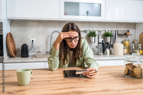 Anxious young woman deciding whether to pay online—holding a credit card and staring at a tablet concerned about fraud, hidden fees, and tight budgeting while shopping from home