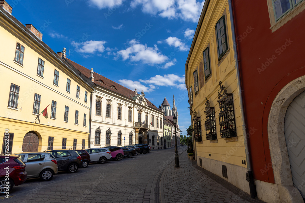 Obraz premium Picturesque European street with colorful historic buildings, parked cars, and bright blue sky with clouds, capturing the charm of old town architecture.
