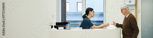 Header of Caucasian senior man interacting with young female nurse at reception desk in medical facility, woman handing document to man, both standing, modern healthcare environment visible