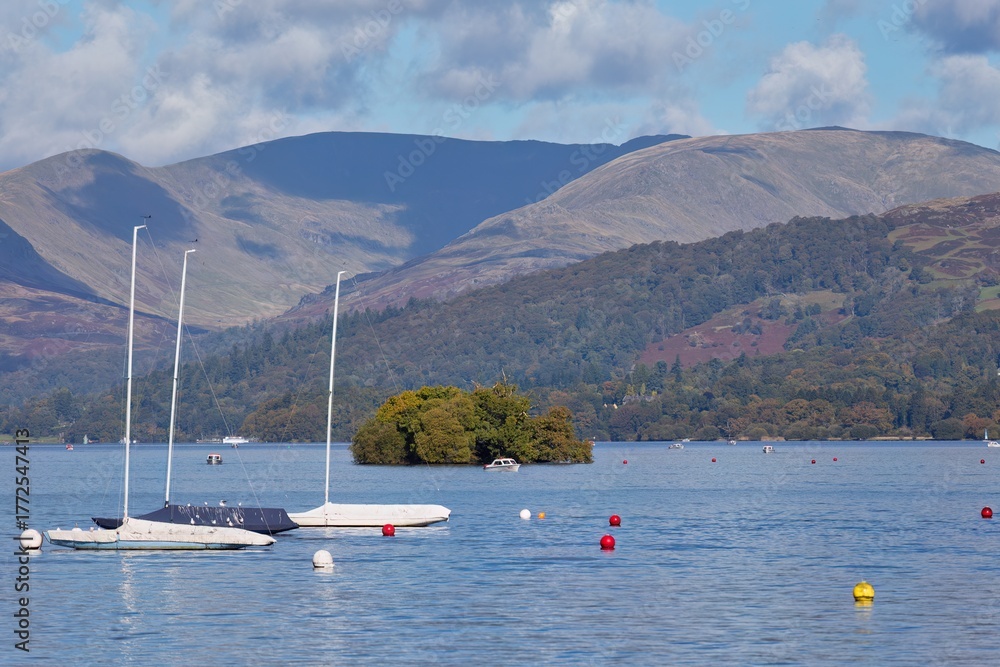 Fototapeta premium Lake scene with sailboats and an island. Mountains rise in the background, and buoys float in the calm water in Bowness-on-Windermere - Lake District - UK