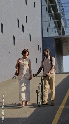Vertical view of middle-aged lady and young man with bicycle walking along paved road after work in business district