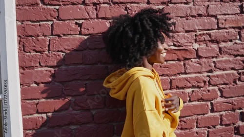 Smiling girl in yellow hoodie stands against red brick wall during sunny day