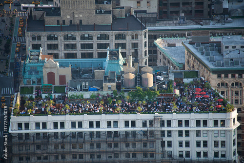 View of a rooftop oasis brimming with people, vibrant greenery, and red umbrellas contrasting against the city's rigid architecture, New York, New York, United States.