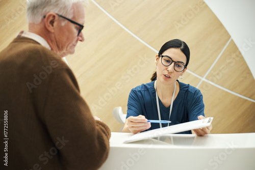 Wallpaper Mural Young female nurse explaining medical documents to senior Caucasian man at reception desk at clinic or hospital, both wearing glasses, healthcare professional assisting patient with paperwork Torontodigital.ca