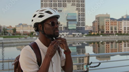Tilt up view of Black young man wearing sunglasses putting on white cycling helmet and fastening it outdoors