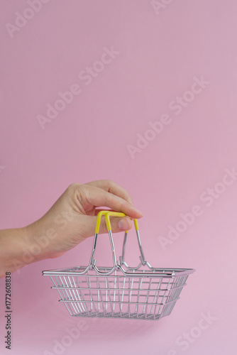 Close-up of a woman's hand holding a metal shopping basket on a pink background. Shopping concept. Space for advertising and text
