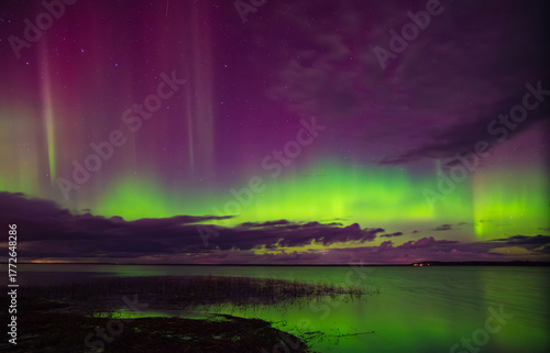 Brilliant aurora borealis in green and purple hues shines above a calm lake and cloudy night sky with faint stars.