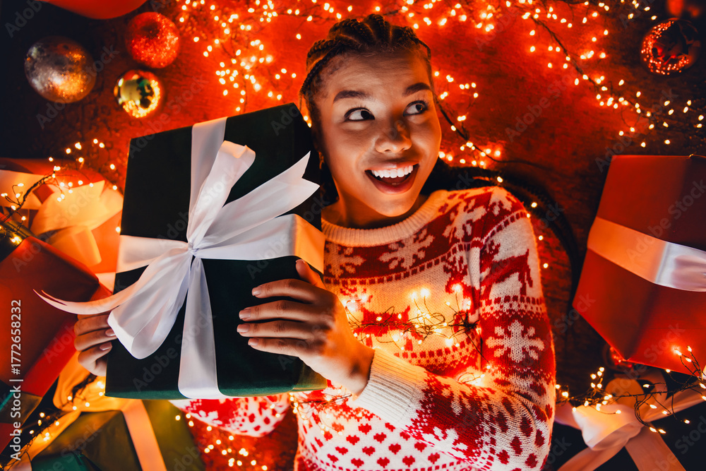 Obraz premium Funny joyful girl holds a large gift box with white ribbon surrounded by Christmas lights in a cozy home celebration