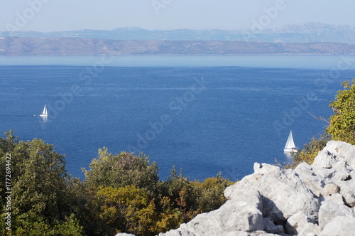 Mediterranean Croatian coastal view with blue sea, white yacht and evergreen trees on rocky hills under bright light. Perfect for travel posters, websites and summer designs.