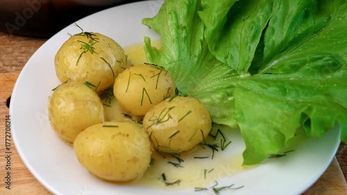 Medium shot: fresh boiled potatoes are being placed on a plate and sprinkled with dill. Slices of ripe tomato are added on a lettuce leaf. A simple and appetizing homemade summer meal.