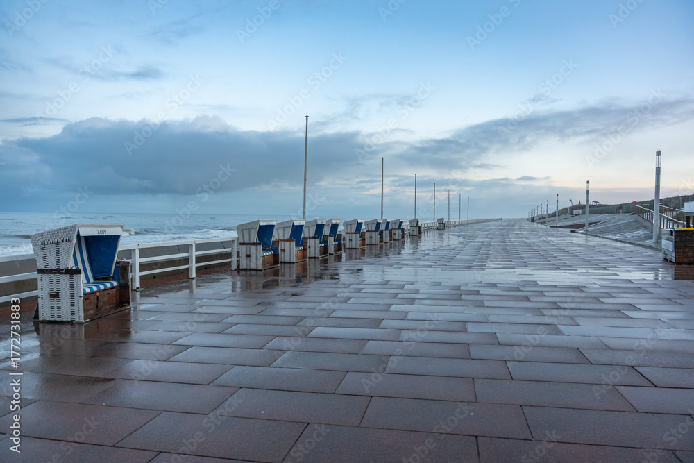 Obraz premium Seaside promenade after rain. Empty promenade with wet tiles and lined beach chairs facing the sea. Calm atmosphere under a brightening cloudy sky.
