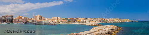 Panoramic Ortigia waterfront in Syracuse, Sicily—pastel historic buildings, marina and boats behind a stone breakwater under a clear Mediterranean sky; serene coastal cityscape.