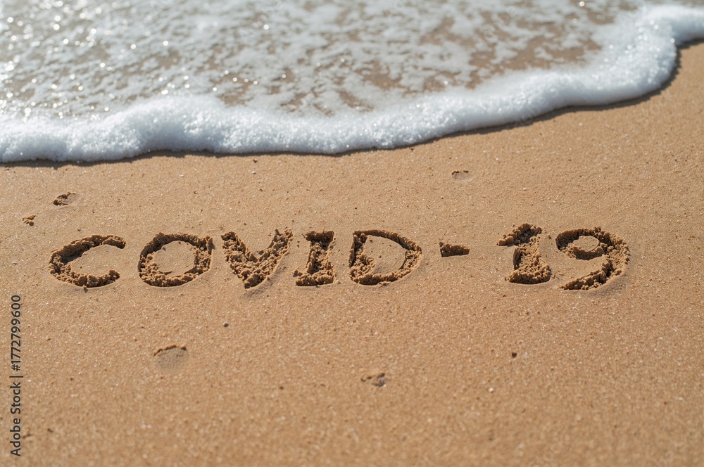 Fototapeta premium Close-up of the phrase covid 19 written in sand on a shoreline. Summer travel limitations caused by outbreaks of respiratory illnesses. Isolation measures in warm regions. Immunization campaigns