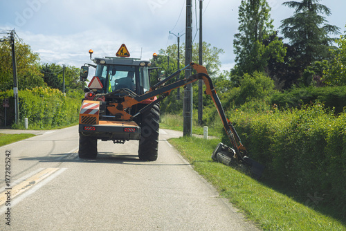 France - A utility tractor with an extended arm attachment trims overgrown roadside vegetation on a sunny day.