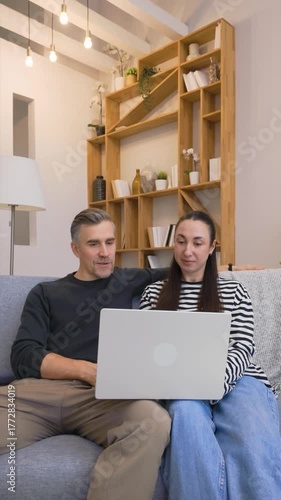 Young couple sitting on sofa at home using laptop together, browsing internet and discussing ideas in cozy bright living room