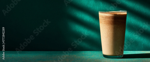 A glass of chocolate milkshake with a layer of cocoa powder on top, placed against a green backdrop