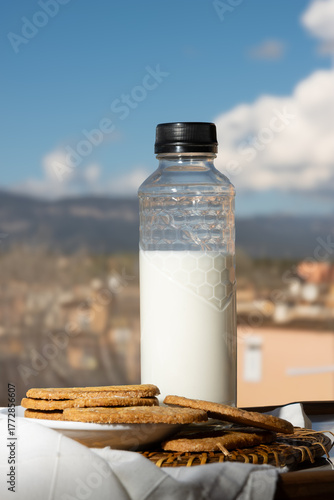Botella de leche y galletas integrales sobre una mesa al aire libre con un fondo de paisaje mediterráneo y cielo azul. 