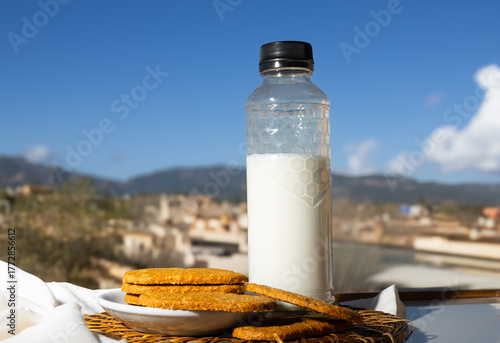 Botella de leche y galletas integrales sobre una mesa al aire libre con un fondo de paisaje mediterráneo y cielo azul. 