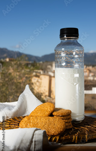 Botella de leche y galletas integrales sobre una mesa al aire libre con un fondo de paisaje mediterráneo y cielo azul. Concepto de desayuno saludable, natural y artesanal en un entorno rural soleado.