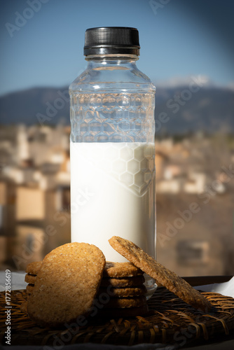 Botella de leche y galletas integrales sobre una mesa al aire libre con un fondo de paisaje mediterráneo y cielo azul. 