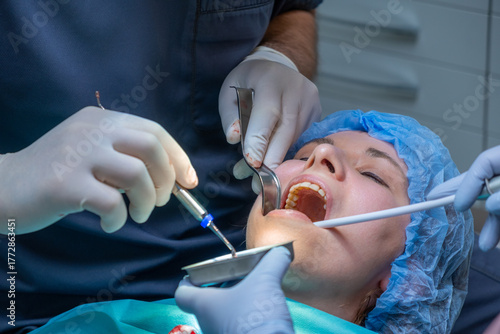 Dental professional works on patient mouth during application of bone graft material. Assistant helps maintain access while woman lying on chair