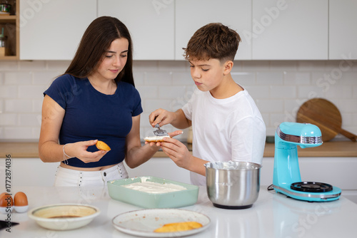 A teenage boy and girl, cooking and spreading cream on ladyfinger biscuits