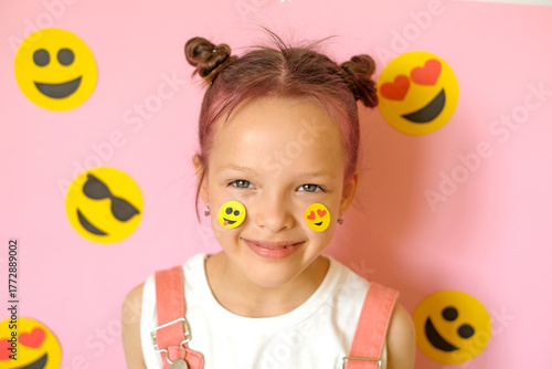 A cute, cheerful girl with emoticons of icons pasted on her cheeks. World Emoji and Social Media Day.