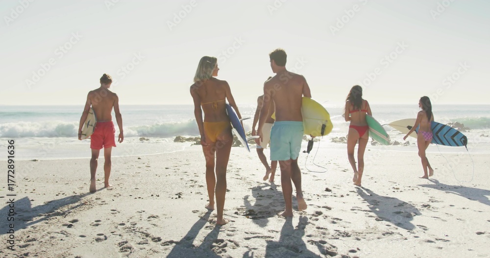 Naklejka premium Walking fit group of six surfers carrying colorful surfboards on sandy beach, showing swimwear