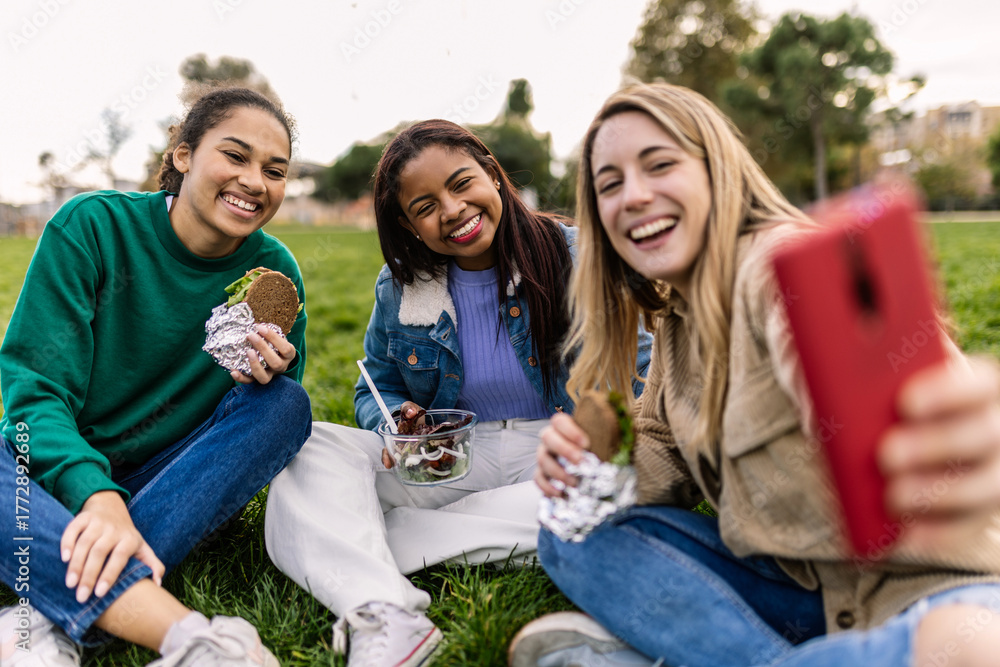 Fototapeta premium Diverse young women friends having social picnic in park, laughing and taking selfie with smartphone, enjoying outdoors lunch