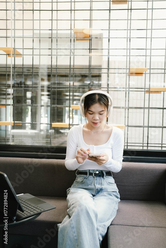 A young woman wearing headphones sits on a sofa, using her smartphone and laptop near a window, enjoying music or working in a relaxed atmosphere. Free Download A young woman wearing headphones sits on a sofa, using her smartphone and laptop near a window, enjoying music or working in a relaxed atmosphere. - GFXLoad