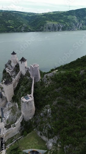 4k vertical footage of Golubac Fortress on the Danube River, Serbia. View of medieval towers, calm water, and wind turbines on the Romanian hills