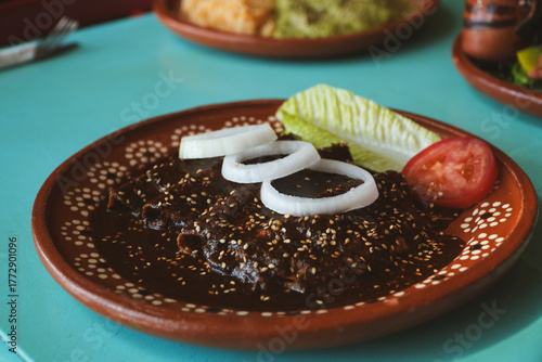 Platillo de enchiladas bañadas en mole, decoradas con rodajas de cebolla, lechuga y jitomate, servidas en plato de barro sobre una mesa turquesa.