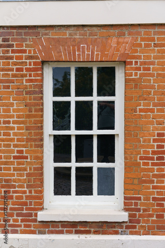 A white-framed window with multiple panes set in a red brick wall, featuring an arched brick lintel with white markings and a lower white section with peeling paint.