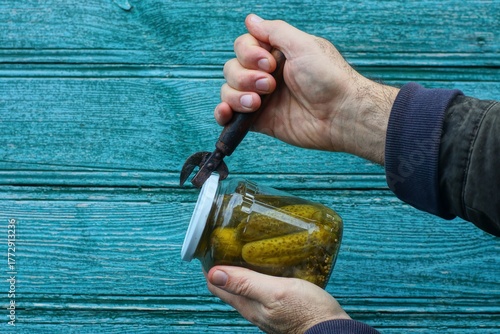 A man opens a small glass jar with salted pickled tasty locally grown green cucumbers with a rusty iron bottle opener with a wooden handle near the wall