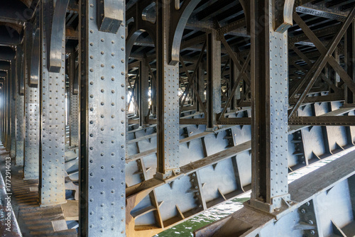 Φωτογραφία A detailed view under a riveted steel bridge, showcasing its robust industrial structure and the bright, rippling water beneath