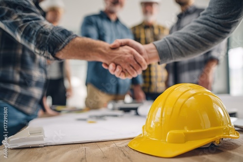 Construction team members shake hands to finalize agreement while wearing safety helmets and gathered around project plans in an office setting