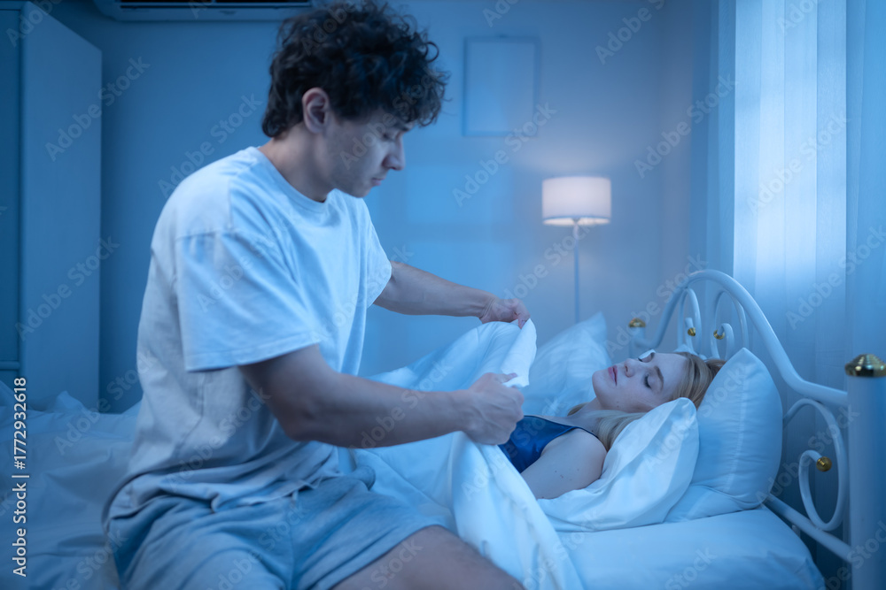 Obraz premium A man sits beside a sleeping woman suffering from a cold in a dimly lit bedroom
